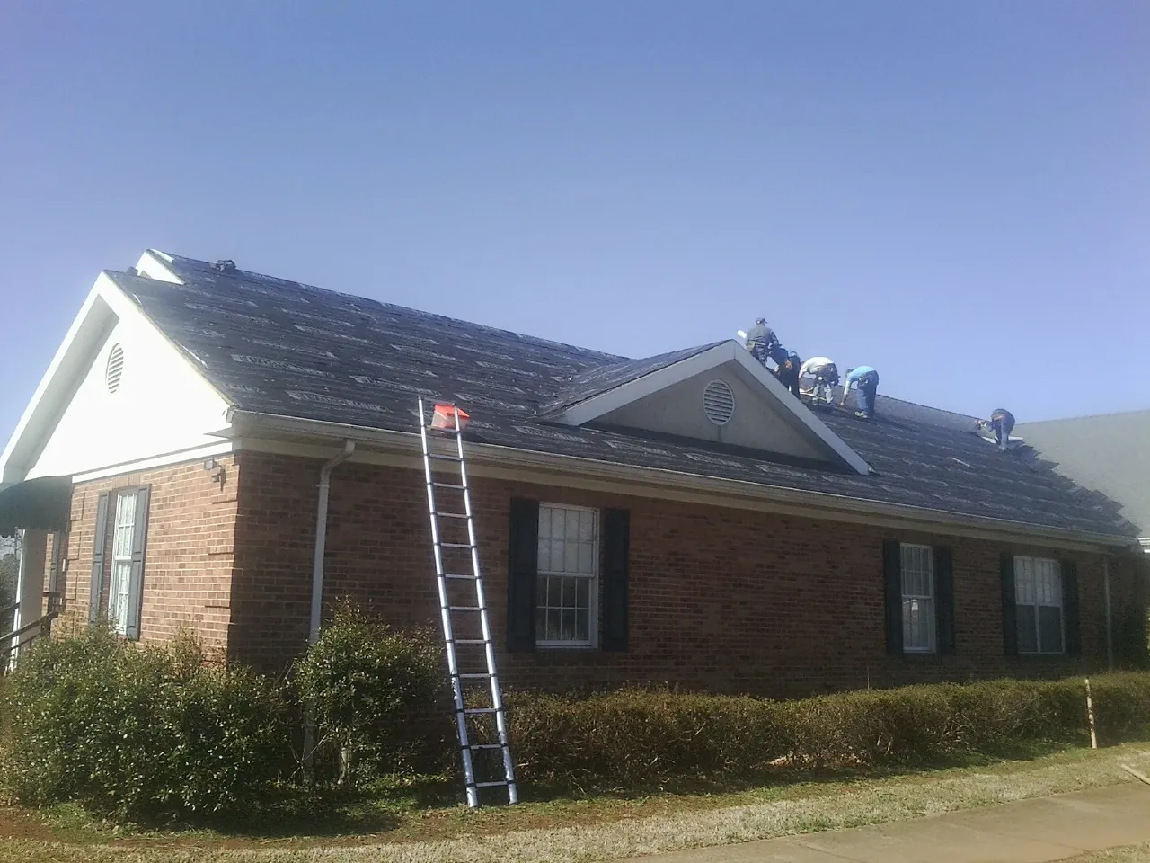 Crew tearing shingles off a brick ranch roof with a ladder set — Emergency Roof Repair work by our crew in Tall Timbers