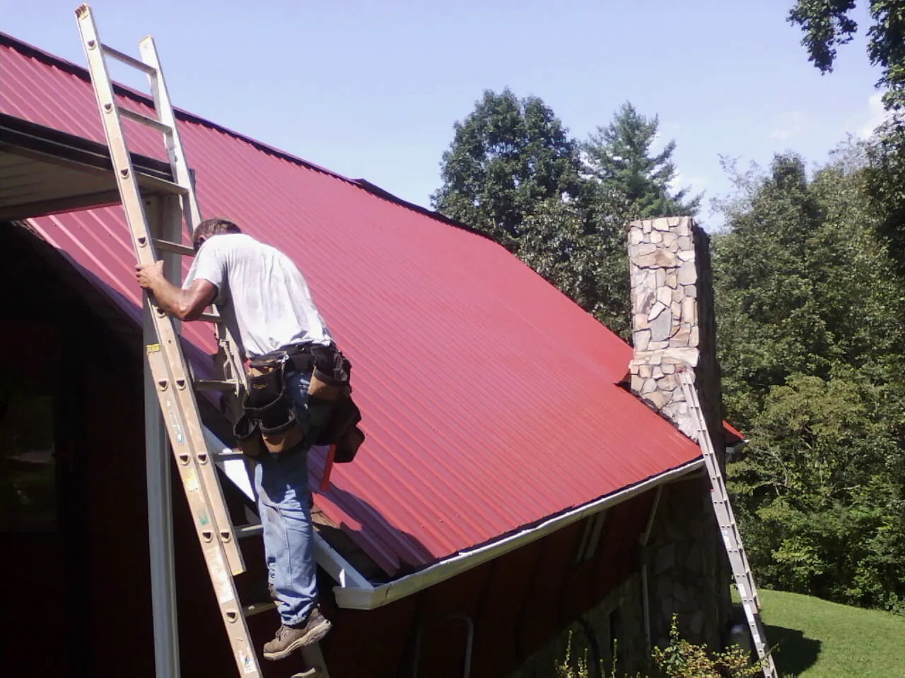 Roofer on a ladder beside a red metal roof and stone chimney — Soffit & Fascia Repair by Roofing Brothers in Tall Timbers